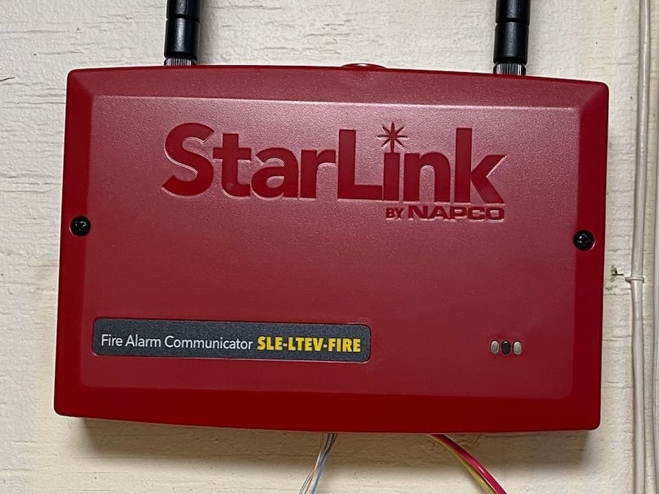 Red cellular fire alarm communicator with two antennas, mounted on a wall above its wiring