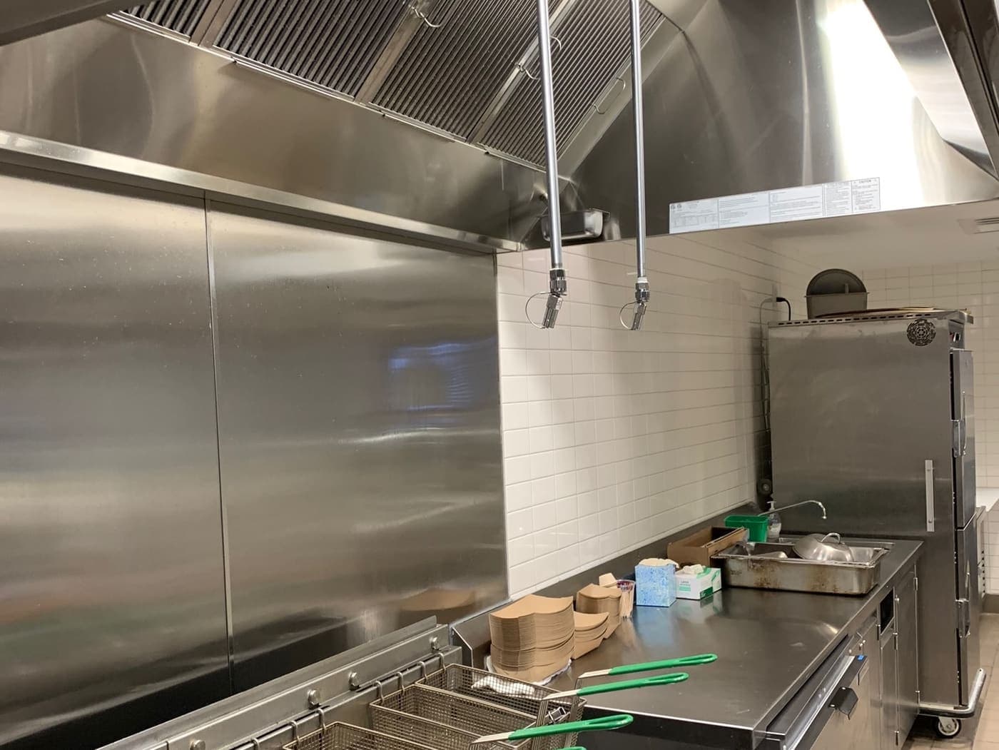 Two wet-chemical suppression nozzles on drop pipes above a commercial fryer bank, beneath a stainless exhaust hood fitted with baffle filters